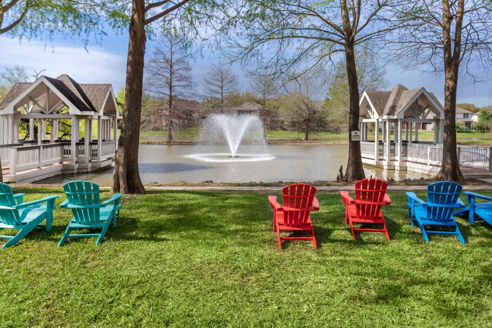Colorful Adirondack chairs arranged on a grassy lawn facing a serene lake with a central fountain and a charming gazebo bridge.