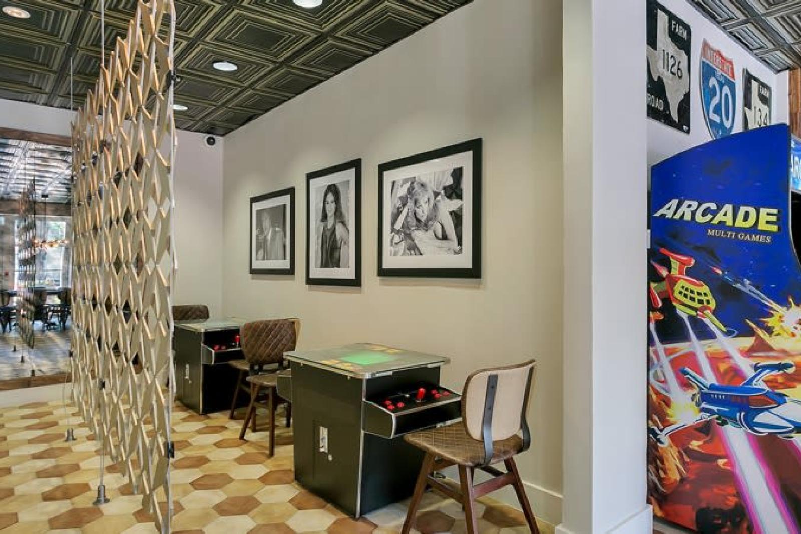 A retro-inspired game room featuring arcade machines, tabletop games, modern chairs, and framed black-and-white portraits on the wall.