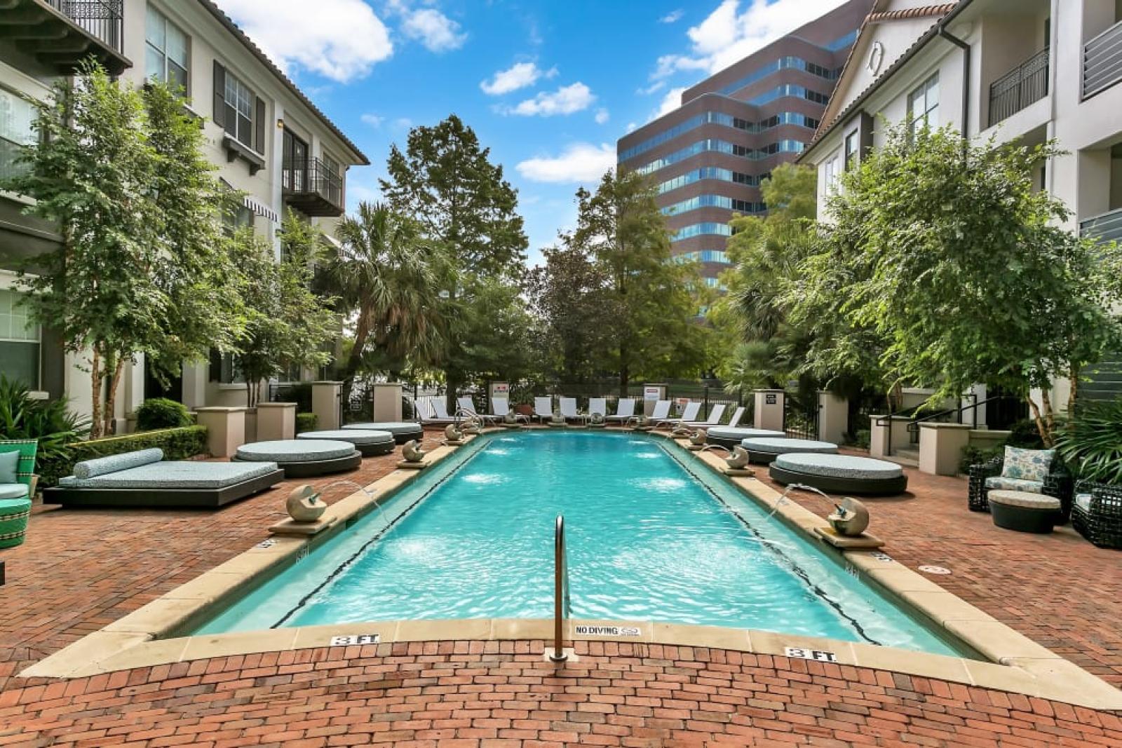 Rectangular pool with cushioned loungers, vibrant landscaping, and modern apartments in the background.