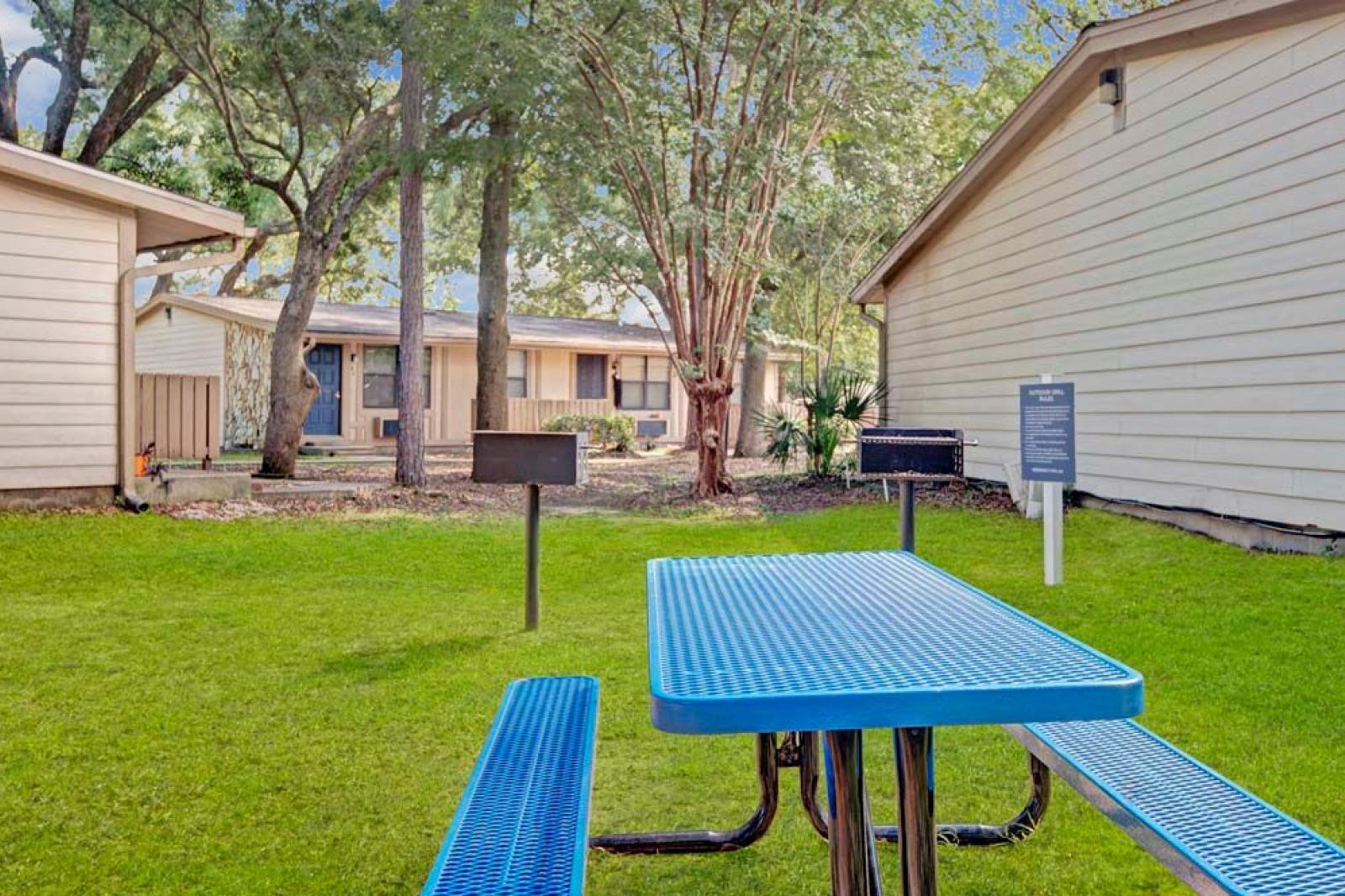 A vibrant outdoor picnic area featuring a blue metal table and a grill, surrounded by lush green grass and mature trees. This image showcases the community amenities and inviting outdoor spaces at Broadview Oaks Apartments.