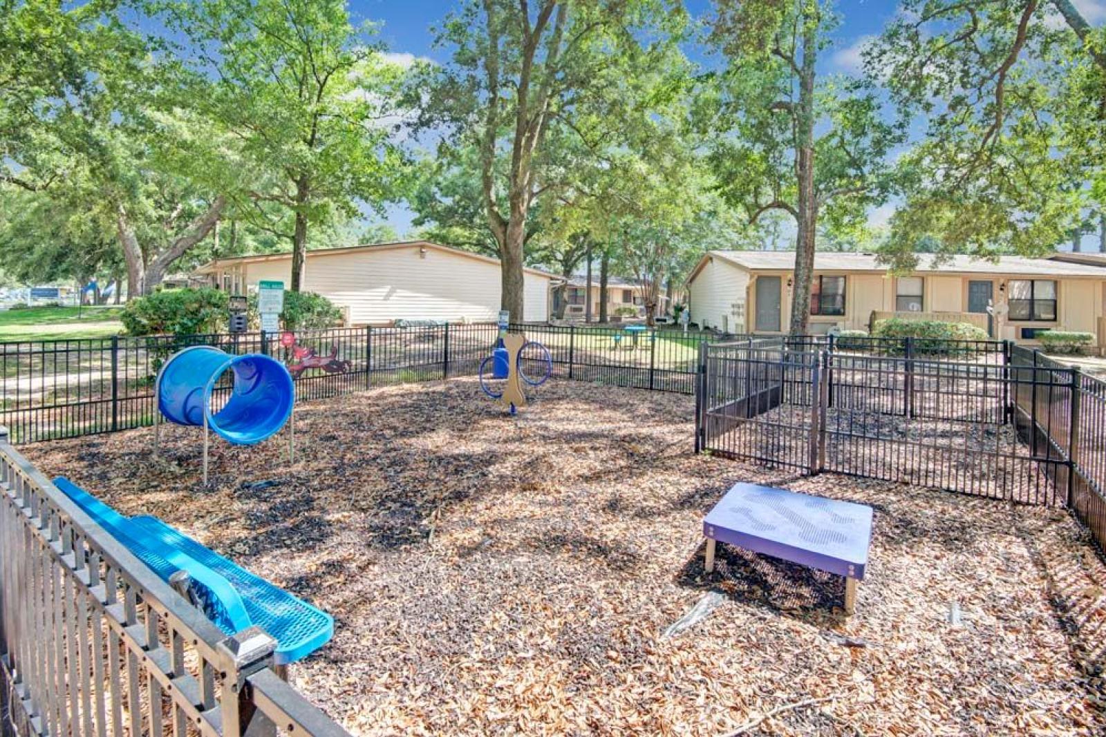 A fenced dog park featuring a blue tunnel slide, climbing equipment, and shaded areas, surrounded by trees and nearby apartment buildings. This image highlights the family-friendly amenities available at Broadview Oaks Apartments.