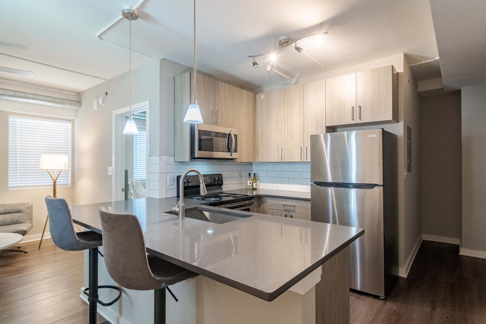 A modern kitchen with sleek stainless steel appliances, including a refrigerator and microwave, set against a light-colored cabinet backdrop. The island features bar stools, and the space is illuminated with stylish pendant lighting