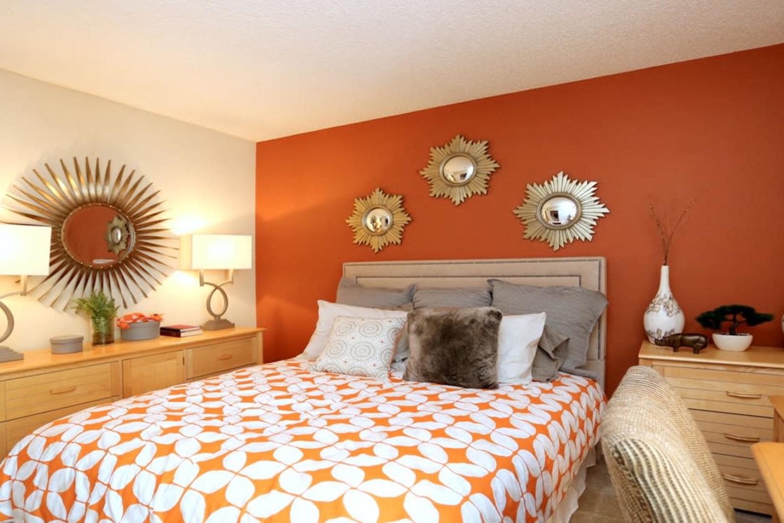  A stylish bedroom at Norcross Crossings Apartments features a bold orange accent wall, elegant decor with sunburst mirrors, and cozy furnishings for a warm and inviting atmosphere.