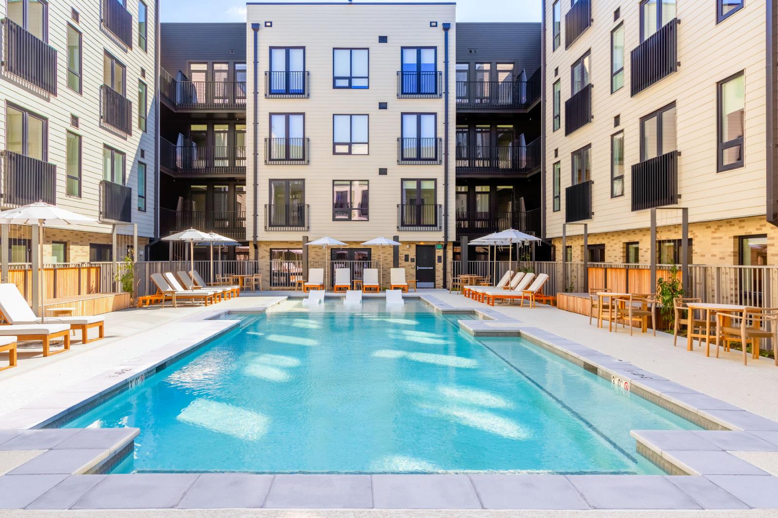 Outdoor pool area at The Edmund Apartments featuring lounge chairs, umbrellas, and in-pool seating surrounded by modern apartment buildings.
