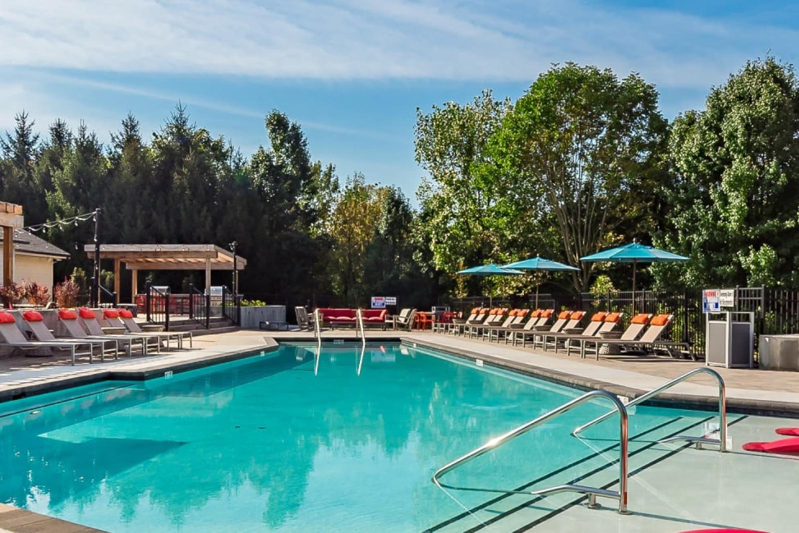 A sparkling outdoor swimming pool surrounded by lounge chairs, umbrellas, and lush greenery, offering a relaxing atmosphere at The Garratt Apartments.