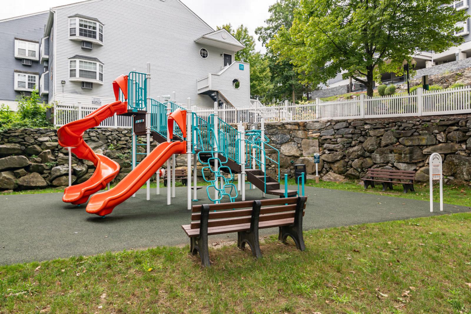 A colorful playground featuring orange slides and turquoise climbing structures, surrounded by benches and set against a backdrop of stone walls and residential buildings.
