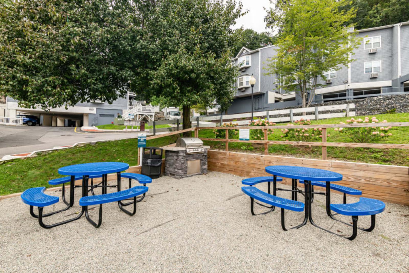 A small outdoor picnic area with blue tables and a barbecue grill surrounded by greenery.