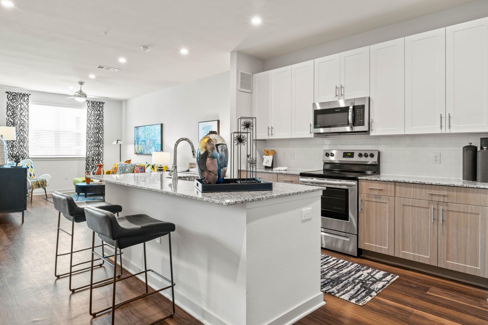 A modern kitchen in Linden Ranch Luxury Apartments featuring granite countertops, stainless steel appliances, and bar seating with stylish black stools. The open-concept design flows into a brightly lit living area with vibrant decor.