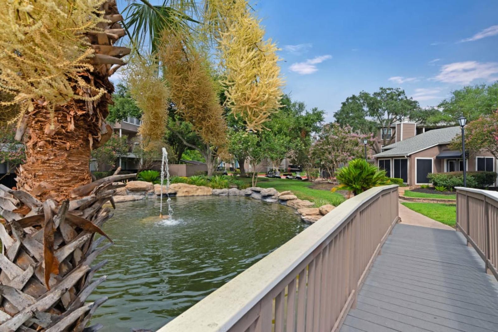 A picturesque outdoor scene with a charming wooden bridge over a tranquil pond, surrounded by lush greenery, blooming flowers, and a decorative fountain.
