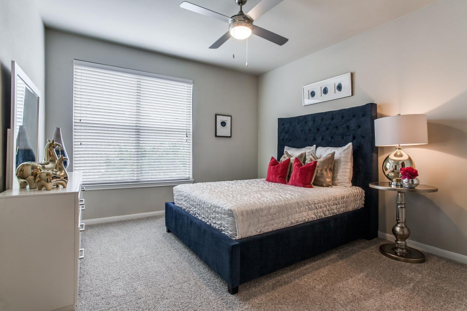 A cozy bedroom with a tufted navy headboard, red accent pillows, and a ceiling fan, creating a relaxing space.