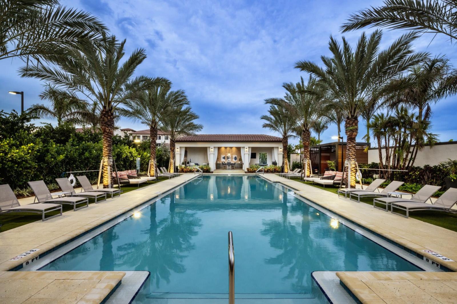 A luxurious swimming pool lined with palm trees and sun loungers, leading to a stylish poolside lounge with cabana seating at Locklyn West Palm Luxury Apartments.