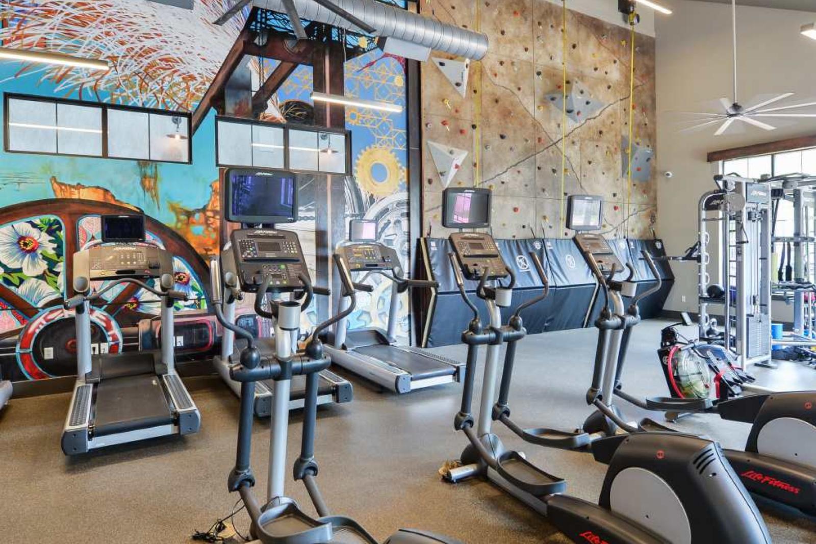 "Modern fitness center at Riverworks Luxury Apartments with treadmills, ellipticals, and vibrant wall art alongside a rock climbing wall."