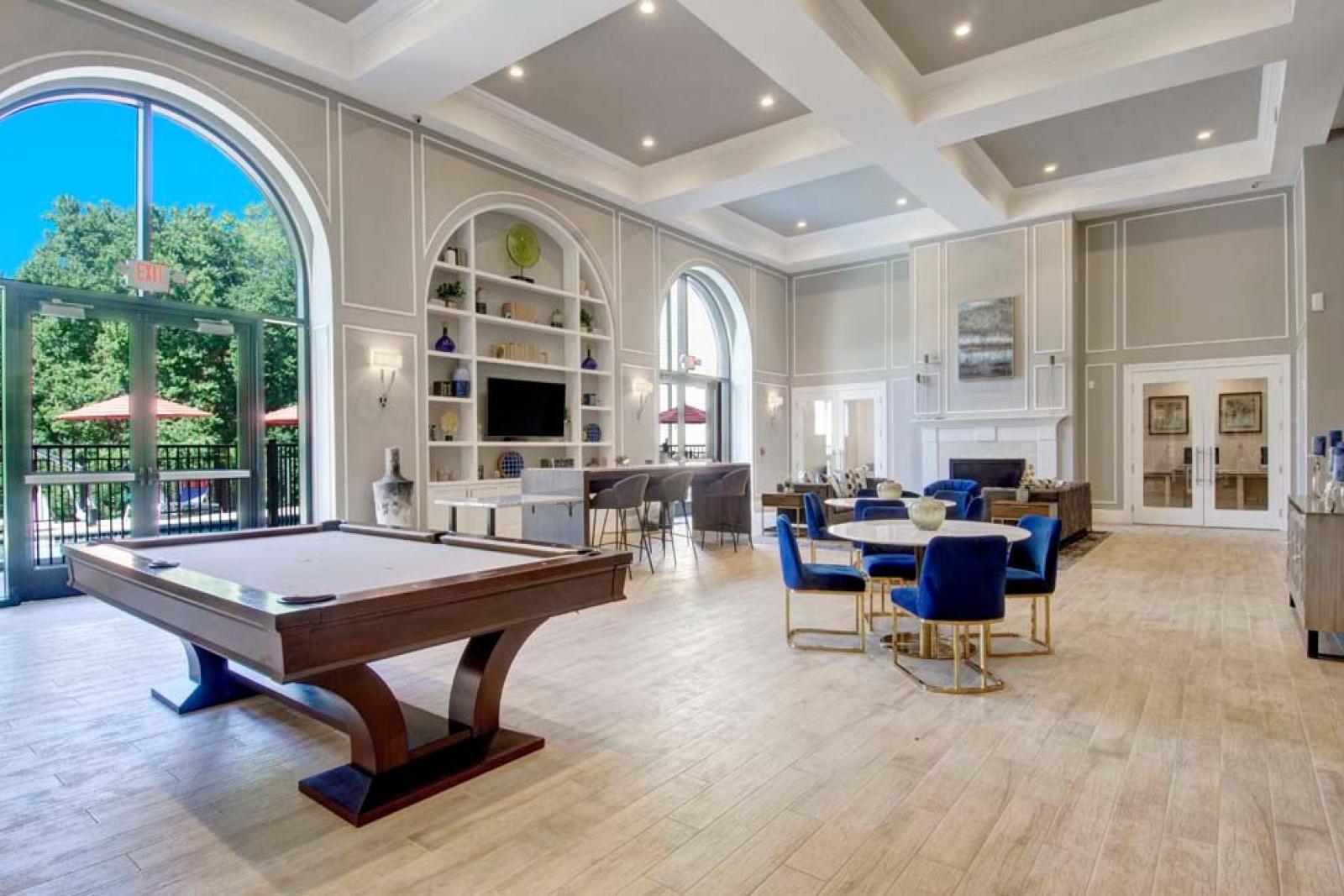 A stylish clubroom with high ceilings, a pool table, and comfortable seating, featuring large windows and access to outdoor spaces.