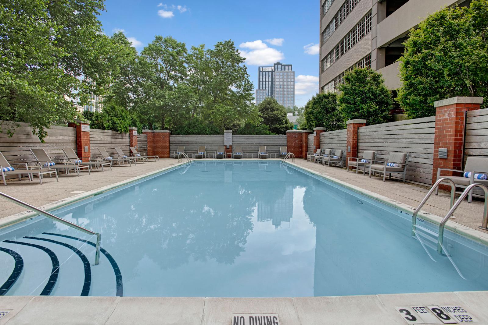 A spacious outdoor pool area with modern seating, trees, and the apartment buildings in the background.