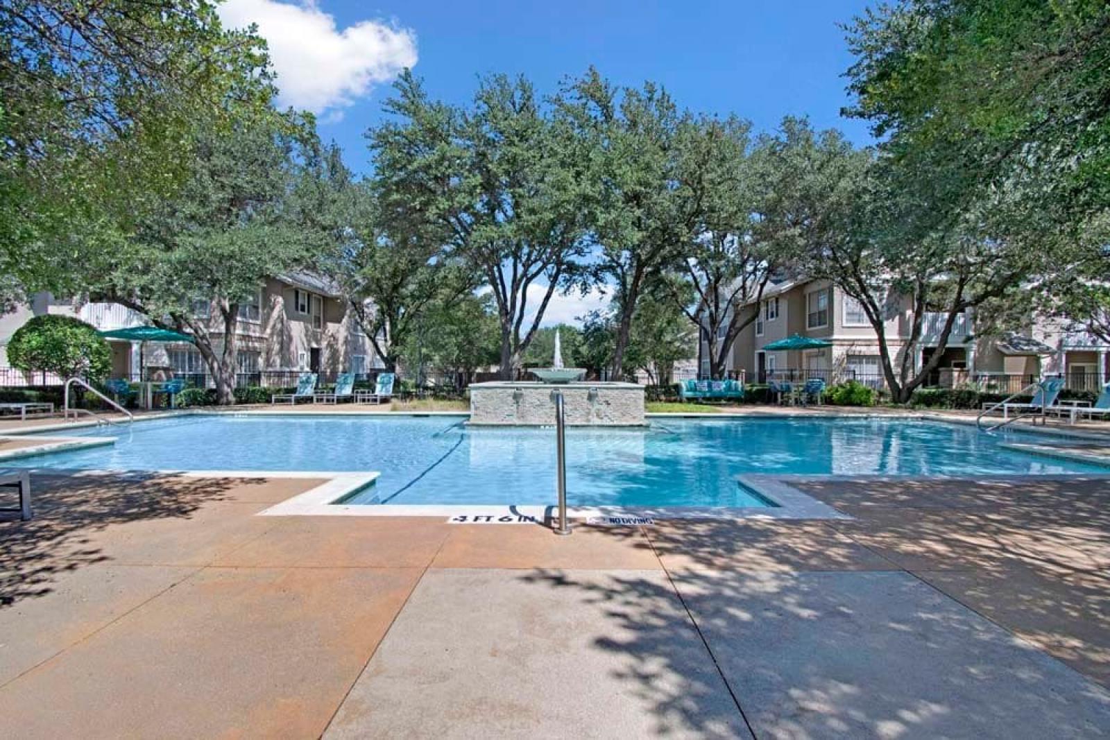 Sparkling pool with a stone water fountain, surrounded by mature trees and sun loungers.