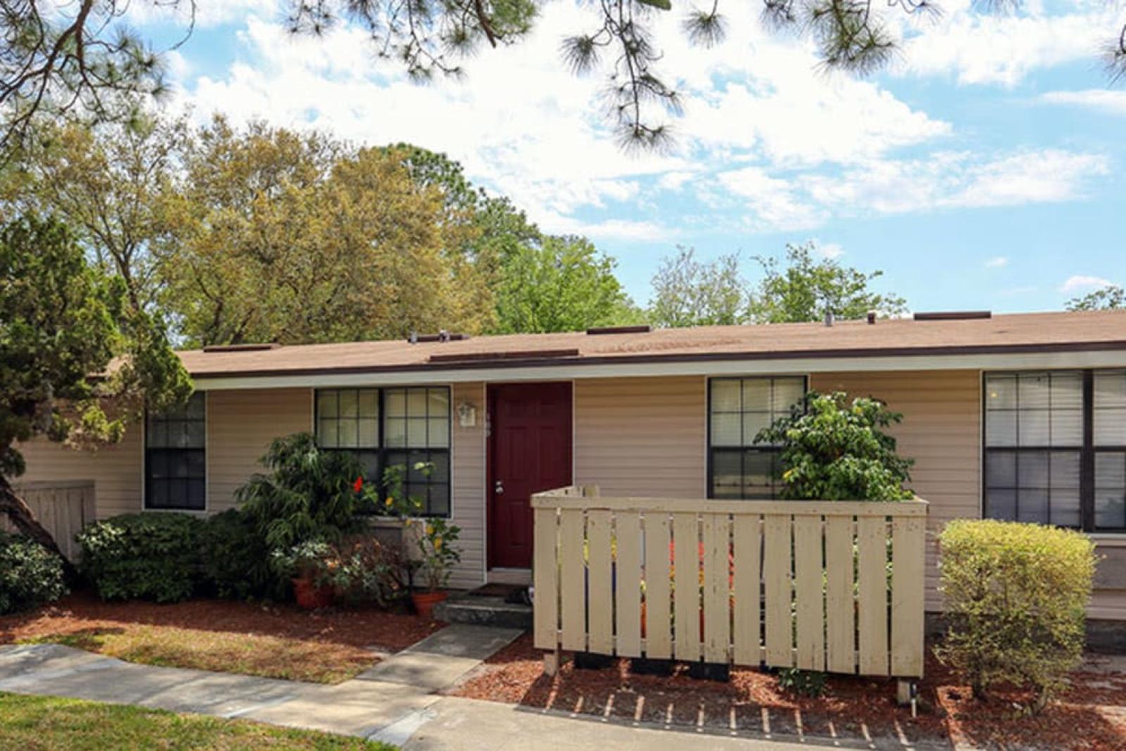 Beige home with a red door, white picket-style fencing, and lush landscaping.