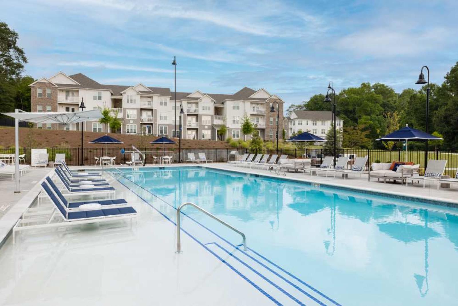 A sparkling outdoor pool surrounded by elegant lounge seating and shaded cabanas, set against a backdrop of luxury apartment buildings and green space.
