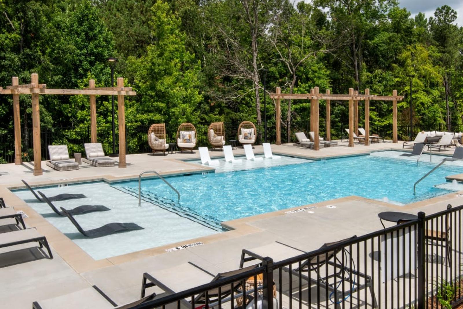 An inviting pool with lounge chairs, surrounded by lush trees and pergolas on a sunny day.