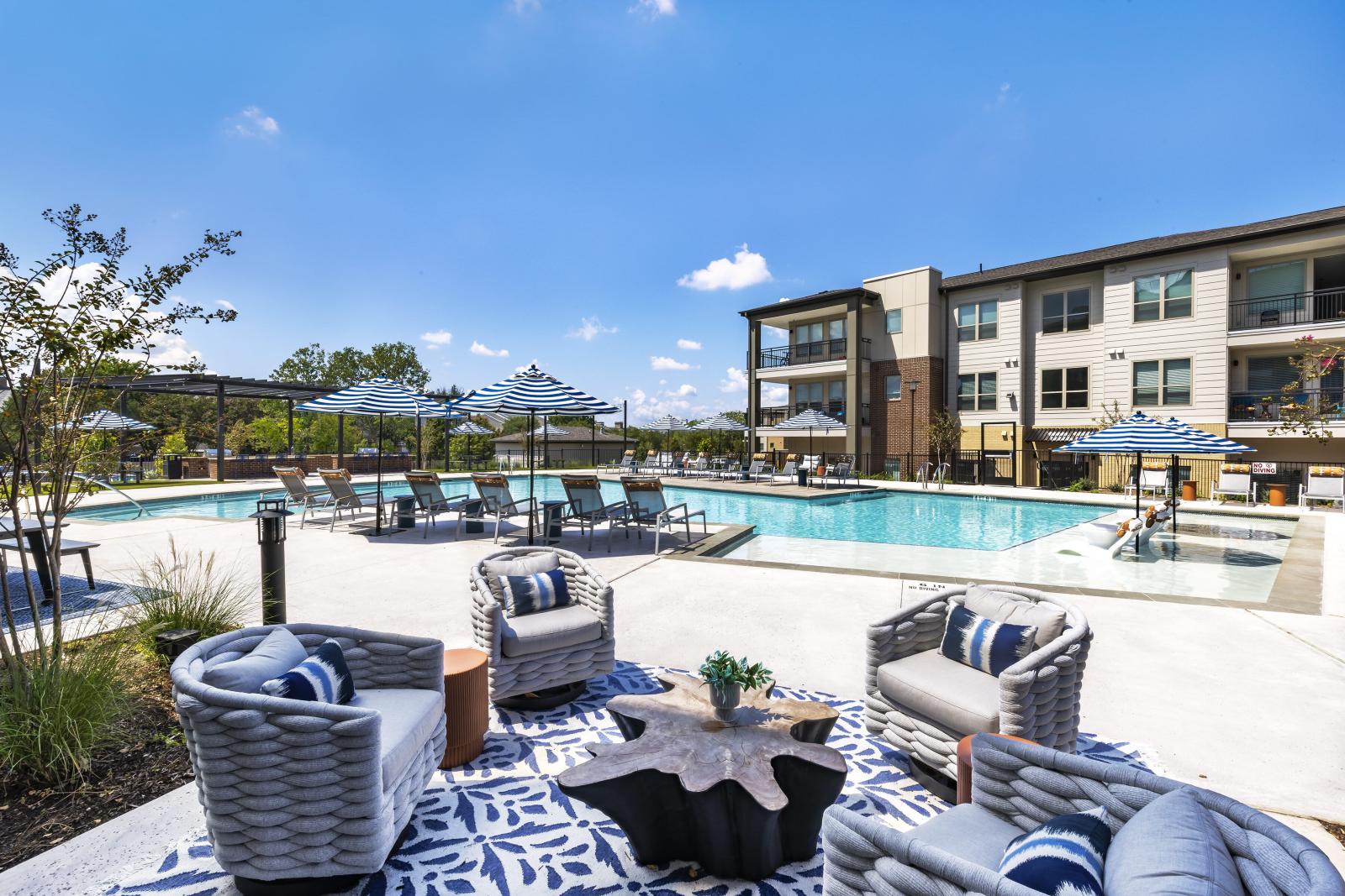 The luxury pool area features stylish woven chairs, a sculptural table, and blue-patterned umbrellas, offering a perfect spot for relaxation.