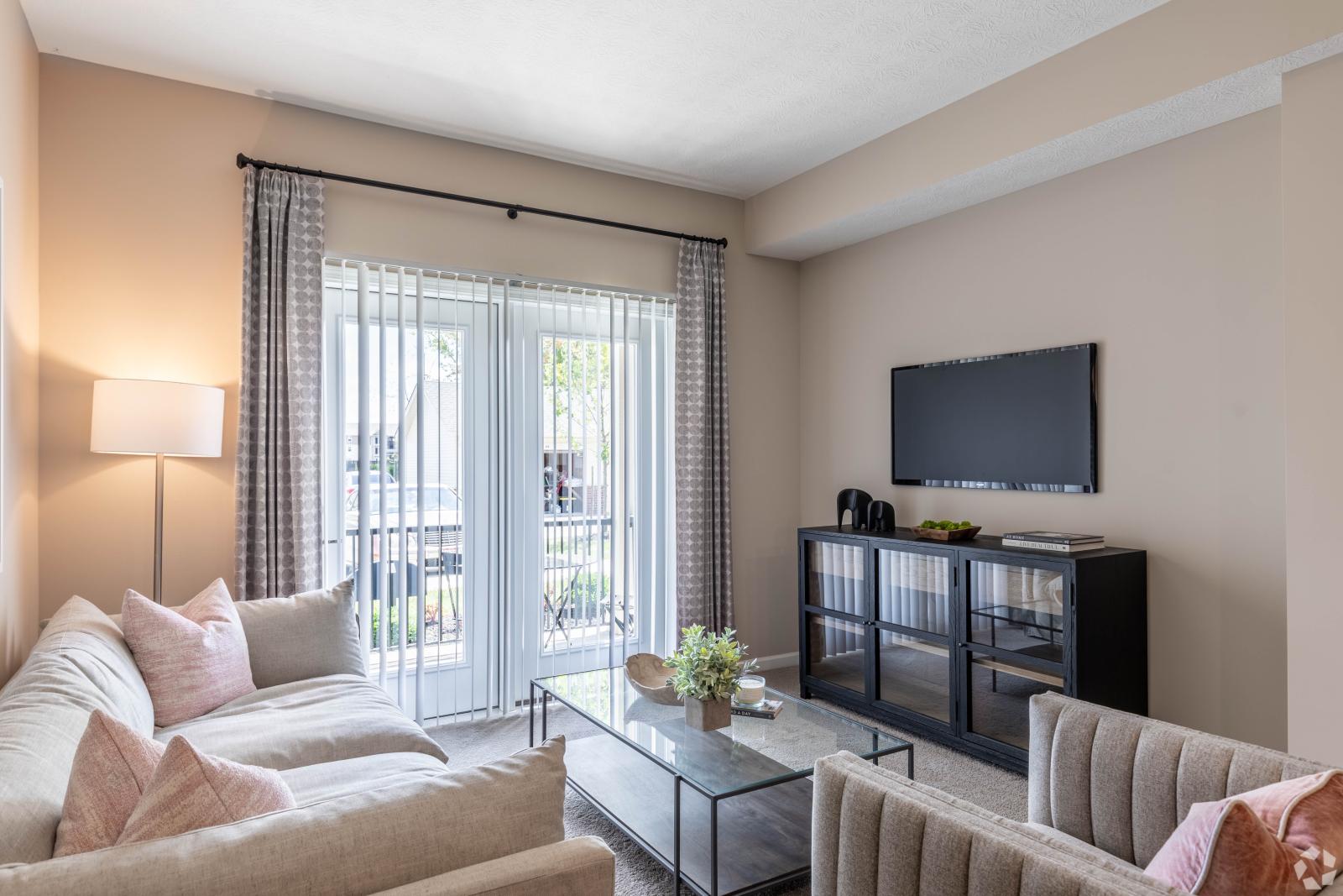 A cozy living room at Victoria Manor Luxury Apartments with soft beige furnishings, large windows, and a wall-mounted TV.