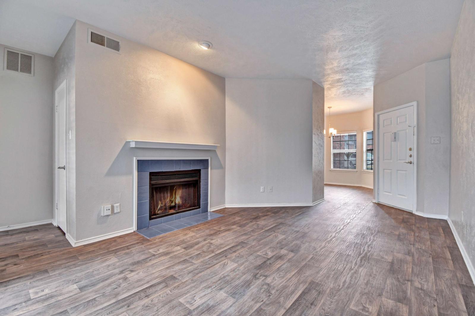 Spacious living area featuring wood-style flooring, a cozy fireplace with a white mantle, and a view into the dining nook near the front entrance.