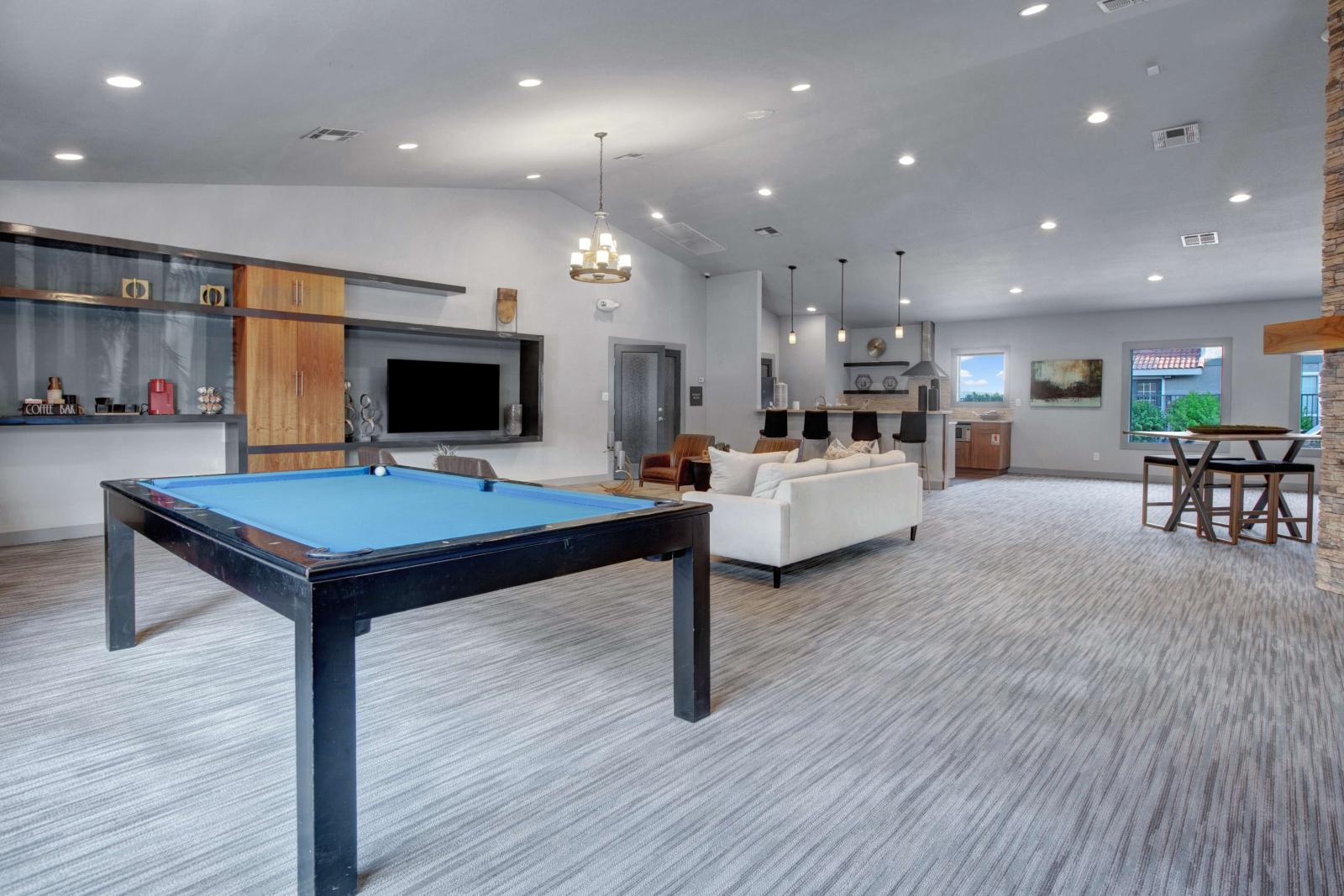 Modern clubhouse interior featuring a blue felt pool table, cozy seating area with a white couch, bar stools at a kitchen counter, and large windows allowing natural light.
