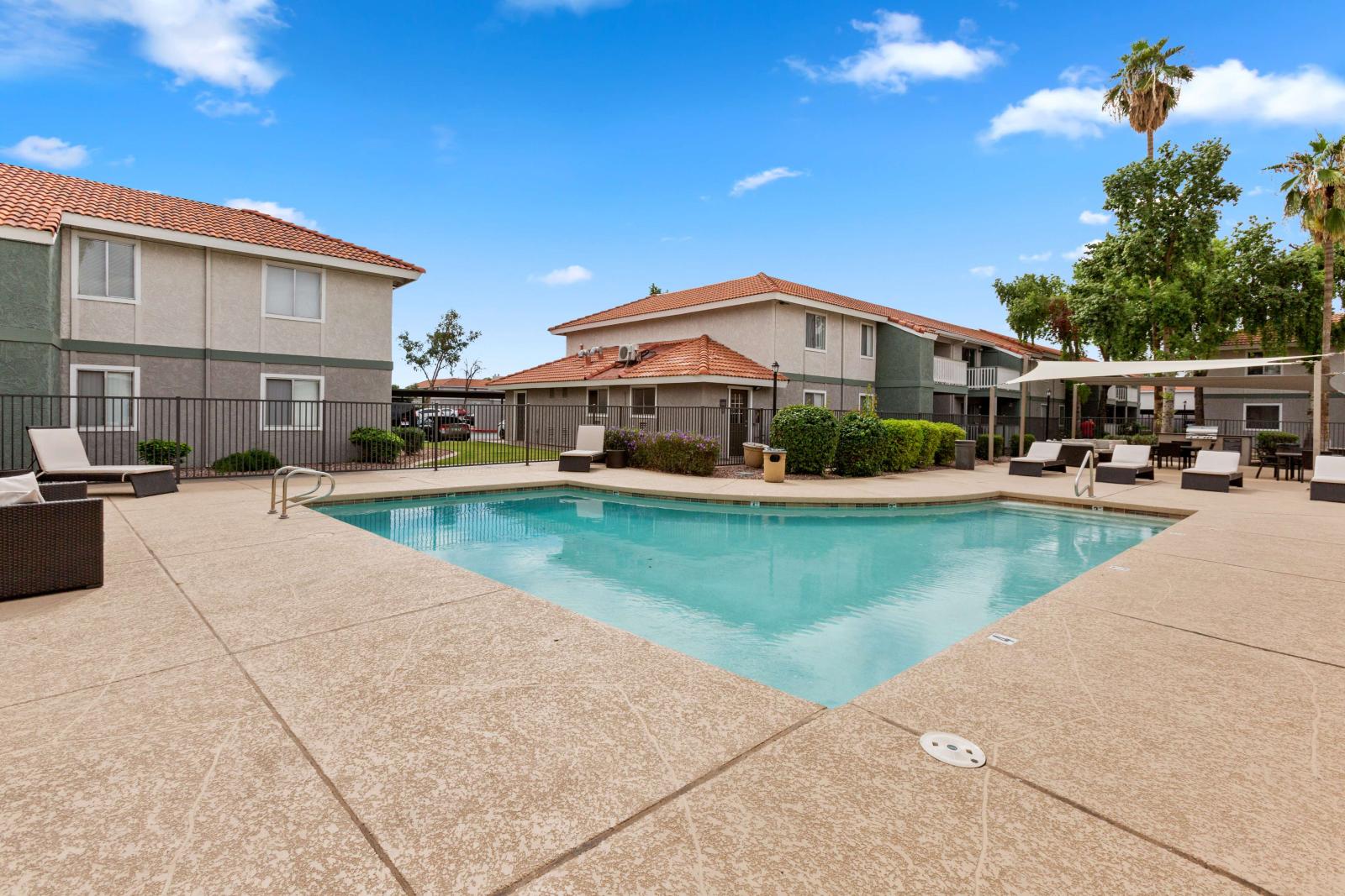 Outdoor swimming pool surrounded by a spacious sundeck with modern lounge chairs, set against a backdrop of two-story apartment buildings and palm trees.