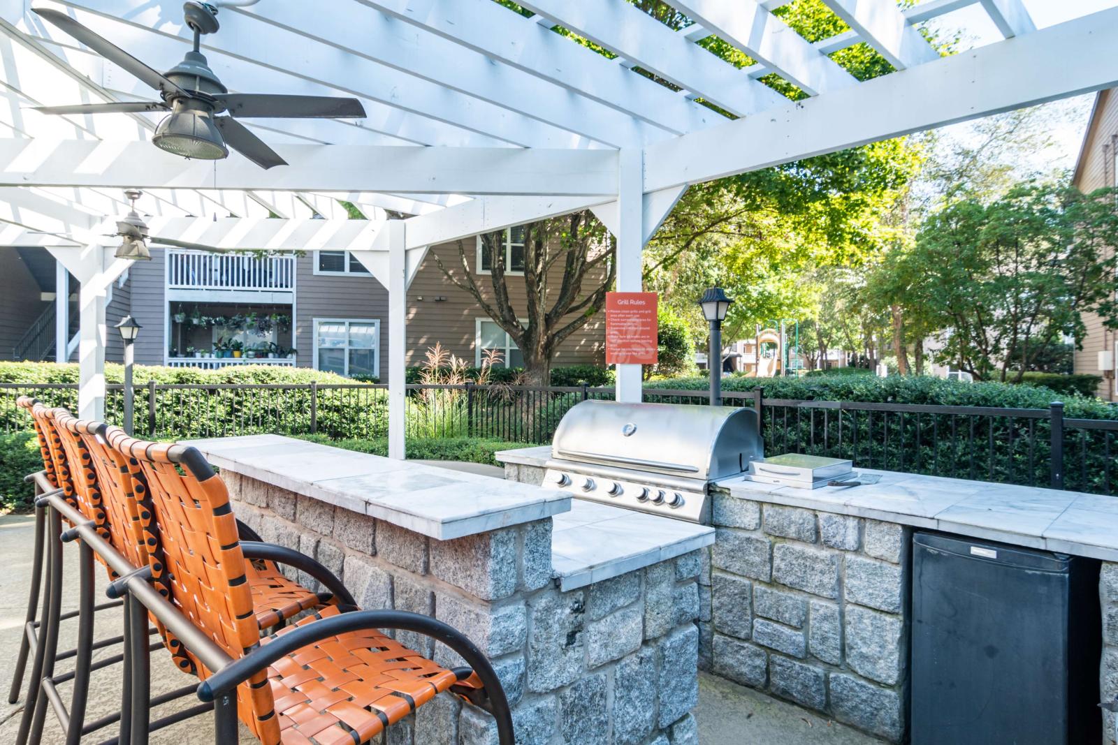 Outdoor grilling station under a white pergola with bar seating and ceiling fans, surrounded by lush greenery.
