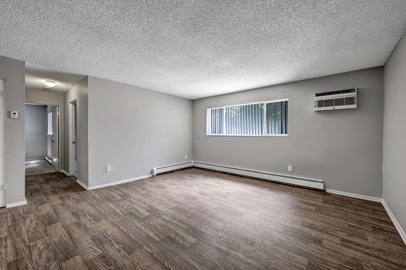 Spacious living room with wood-style flooring, neutral walls, and a large horizontal window with vertical blinds and an in-wall AC unit.
