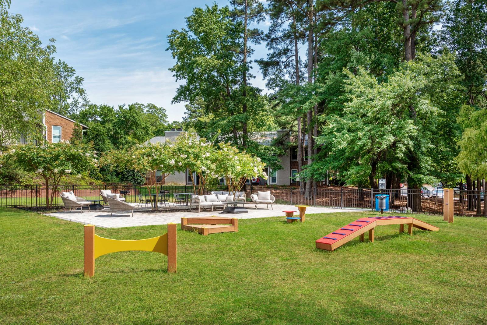 Scenic outdoor pet park and seating area with agility equipment, shaded lounge furniture, and mature trees providing a peaceful, community-friendly space.