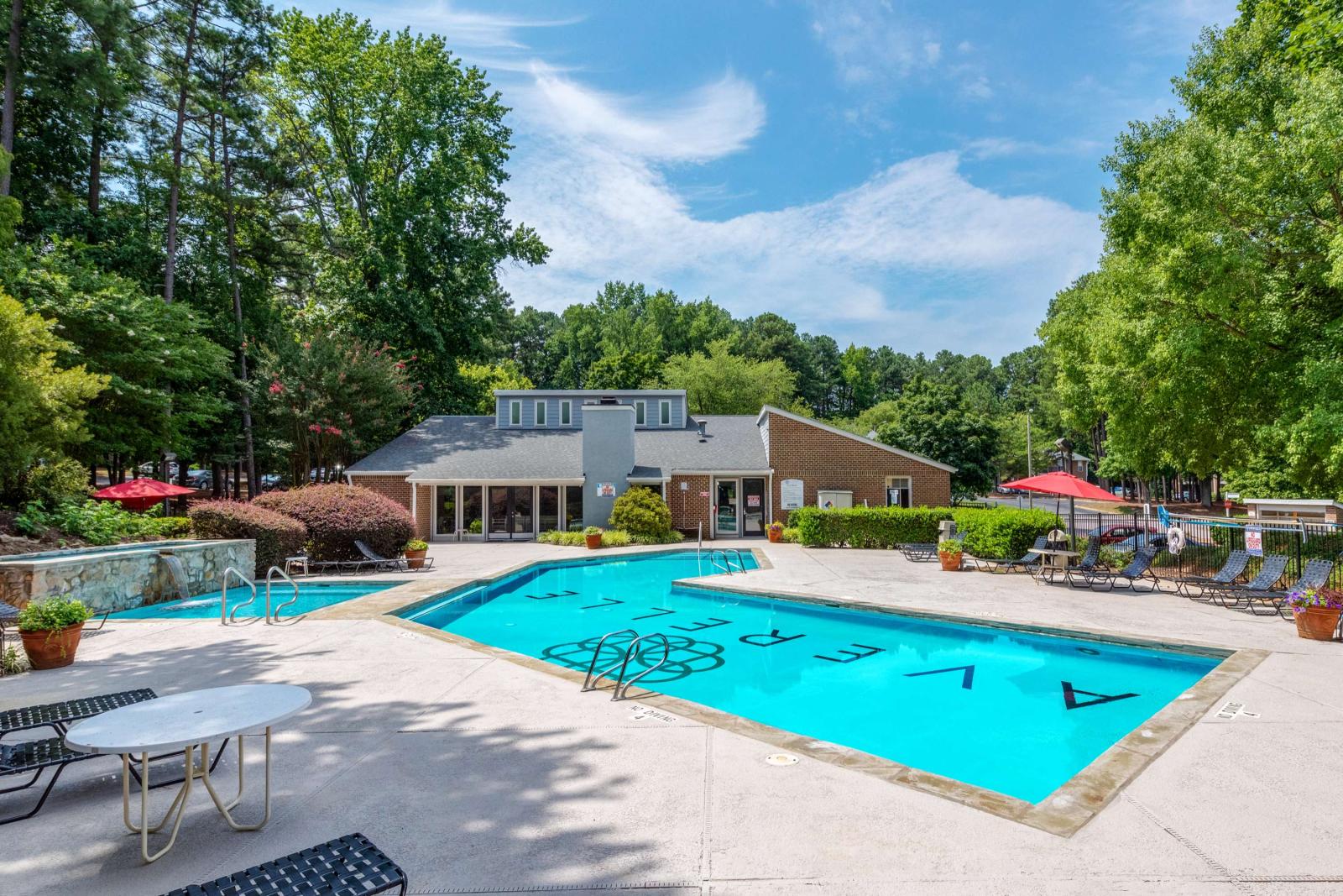 Expansive outdoor pool with crystal-clear water, surrounded by greenery and a brick clubhouse, with plenty of lounge seating and red umbrellas for shaded relaxation.