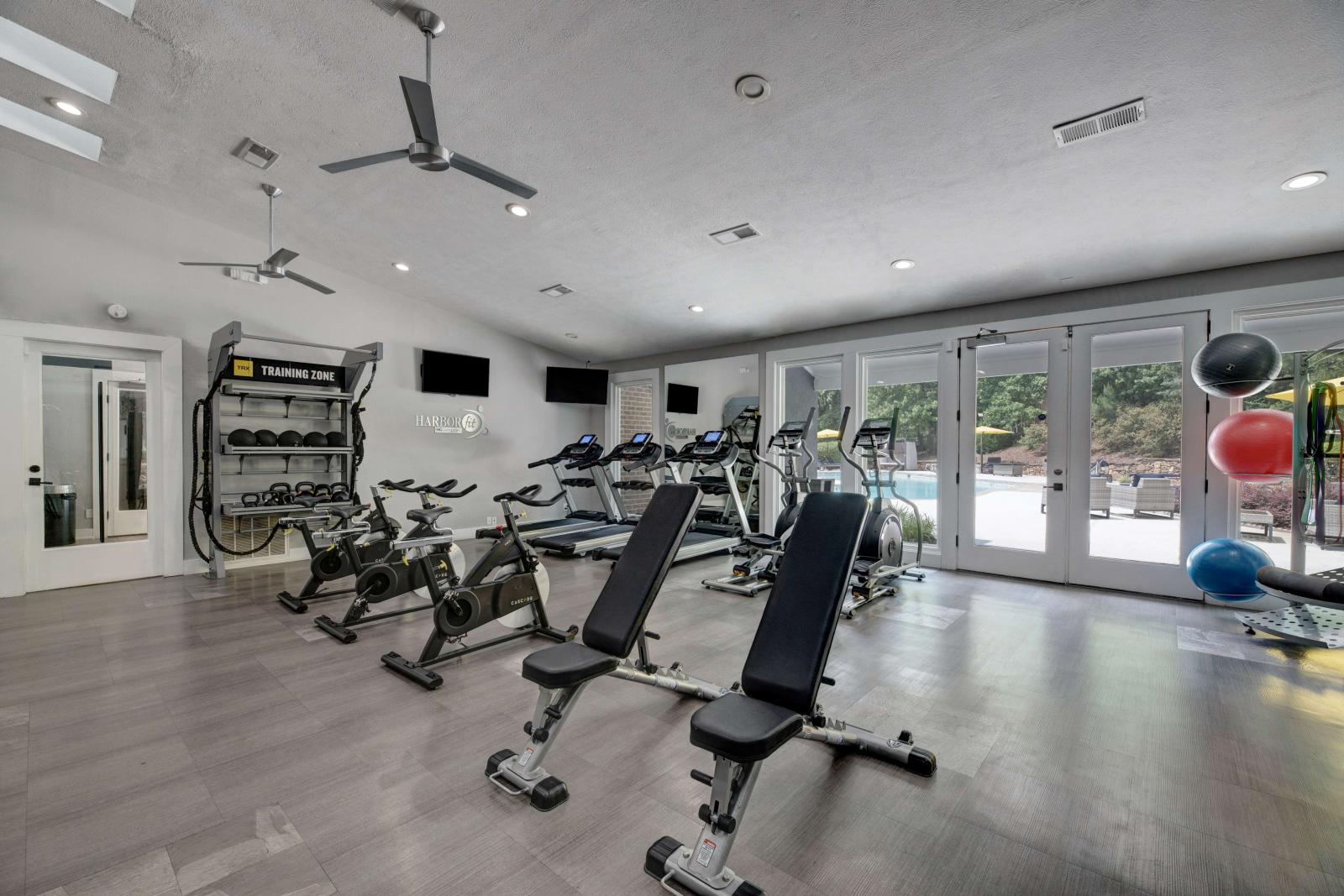 Well-equipped fitness center with treadmills, ellipticals, spin bikes, free weights, and functional training equipment, featuring modern flooring and large windows overlooking the pool area.