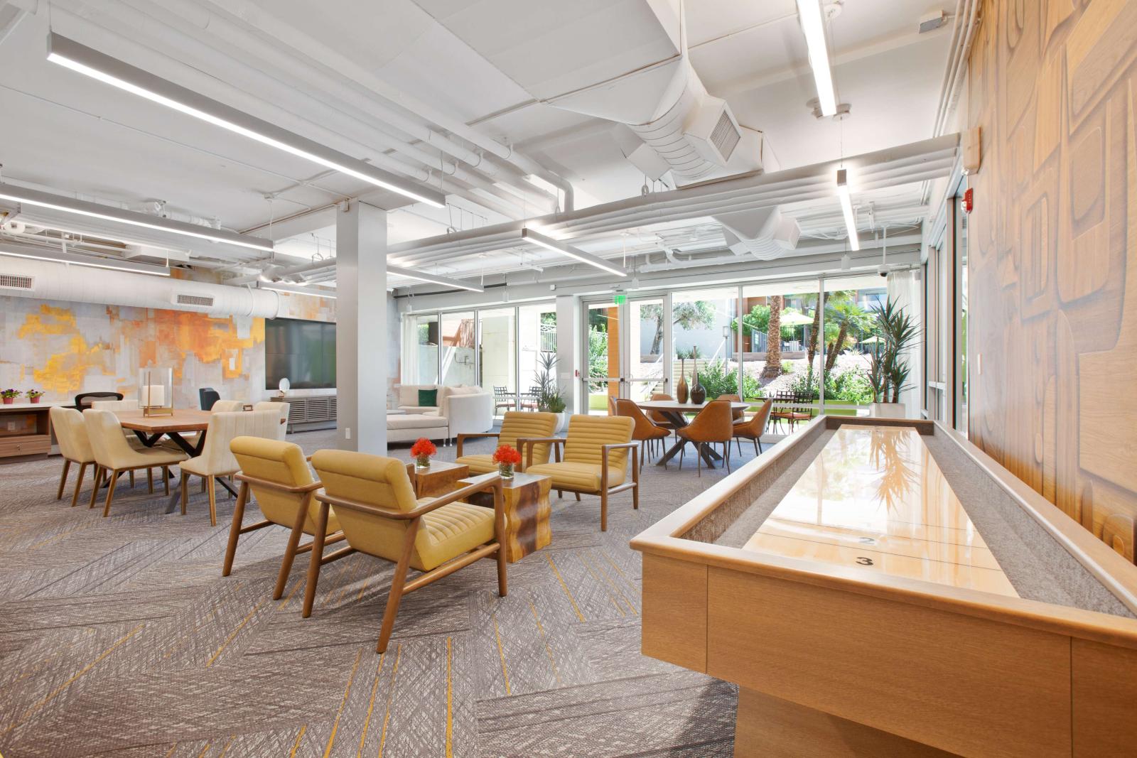 Vibrant resident lounge with a shuffleboard table, colorful accent wall, modern seating areas, and floor-to-ceiling windows that open to a lush outdoor courtyard.