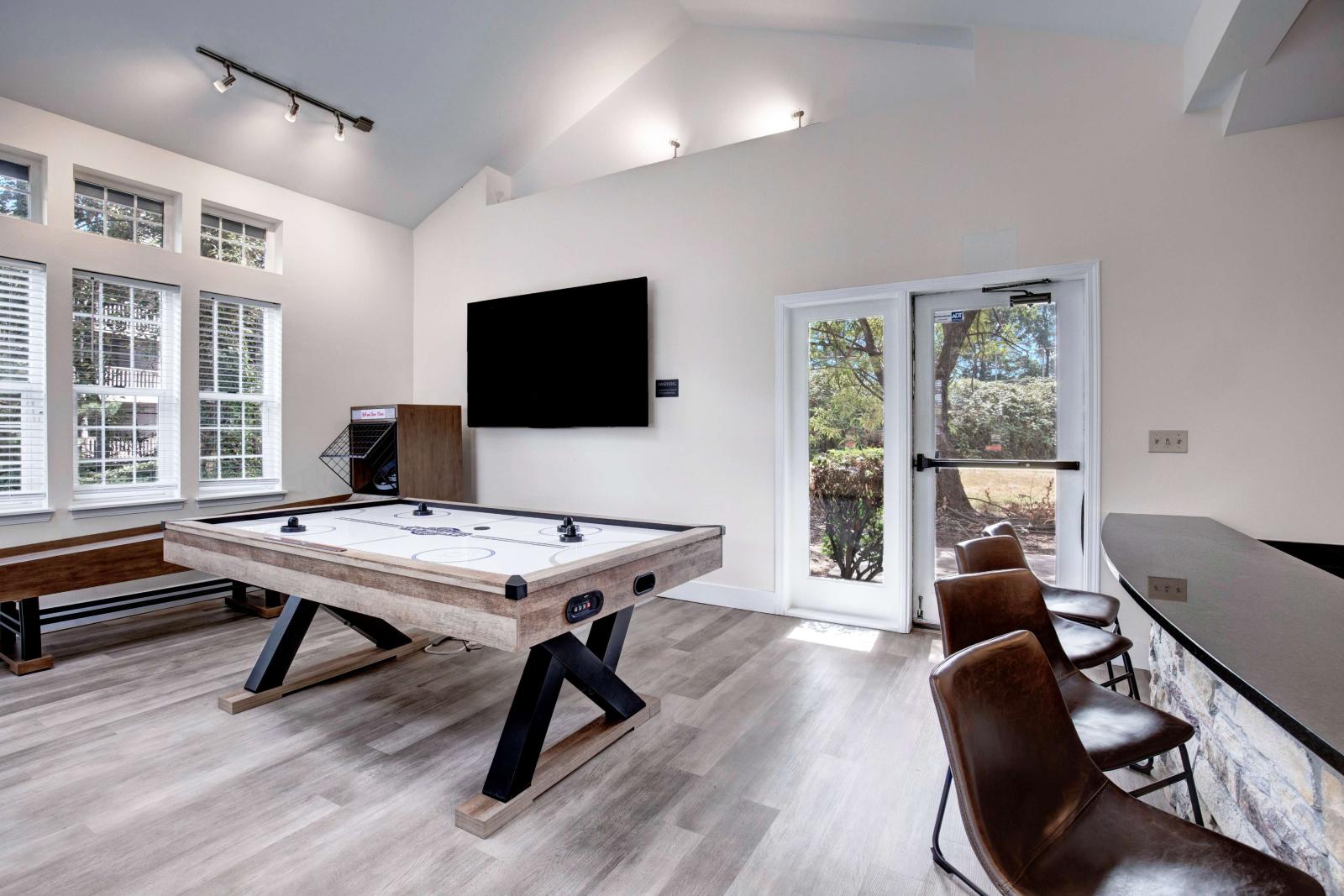 Modern resident lounge with a rustic-style air hockey table, mounted flat-screen TV, shuffleboard, bar seating, and large windows providing natural light.