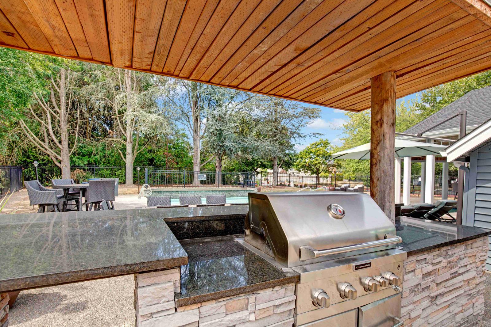 Outdoor grilling station with a built-in stainless steel KitchenAid grill, granite countertops, and a stone base under a wooden pergola. In the background, there is a pool area, patio seating with wicker furniture, and lush trees providing a serene setting.