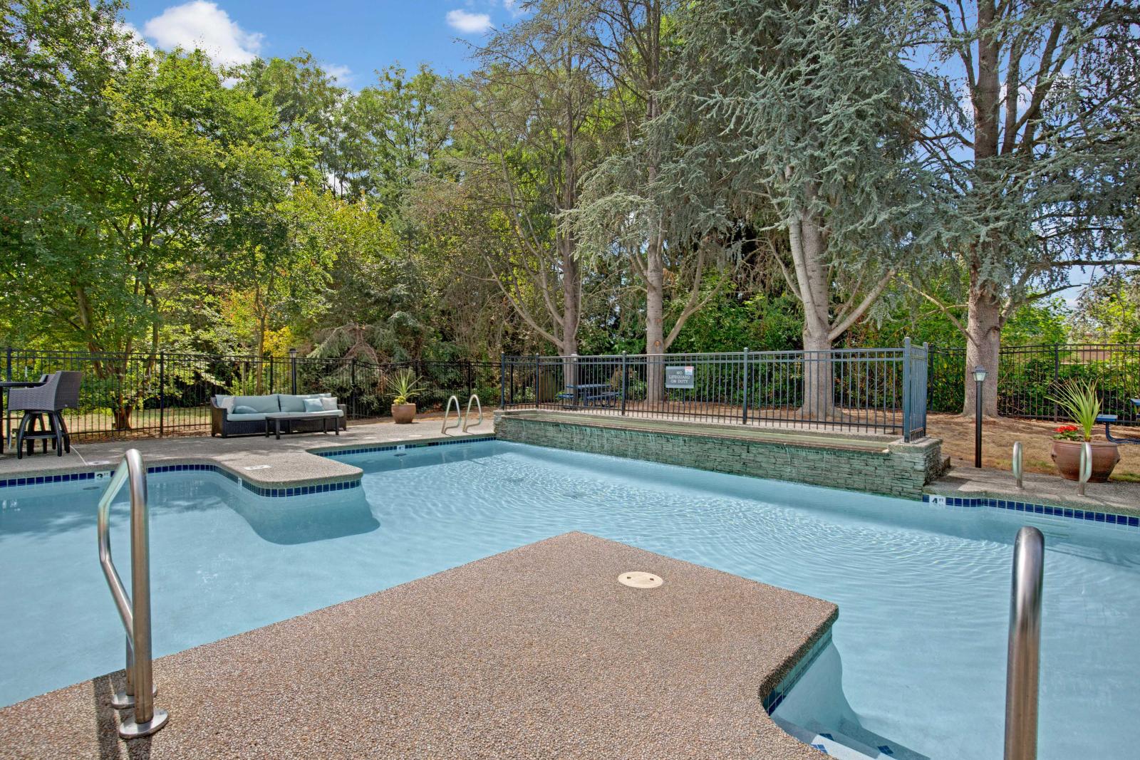 Peaceful outdoor pool surrounded by mature trees and lush greenery, featuring a textured deck area, built-in lounge seating, and raised stone wall for added privacy and charm.