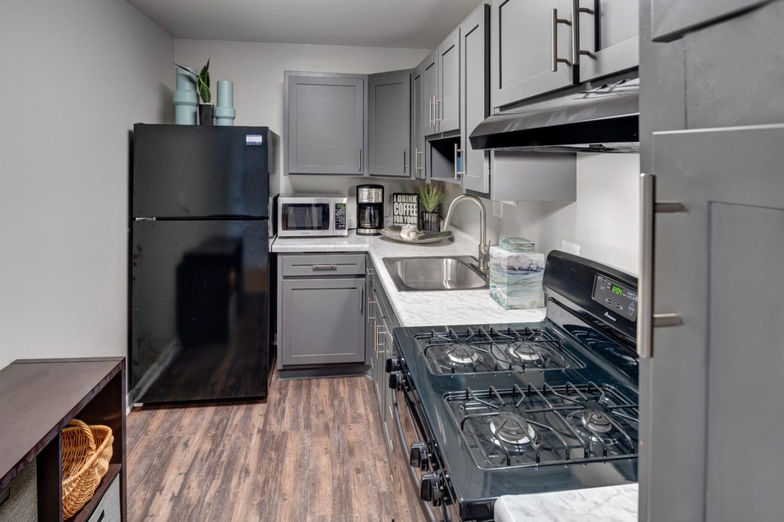 A sleek and functional kitchen at East Broad Plaza featuring gray cabinetry with brushed nickel hardware, white marble-style countertops, and black appliances, including a refrigerator, gas stove, and range hood. The compact kitchen layout is efficiently designed with a deep stainless steel sink and modern fixtures. Decorative elements like a coffee sign, potted plant, and accent jars add warmth and character to the space. ​​