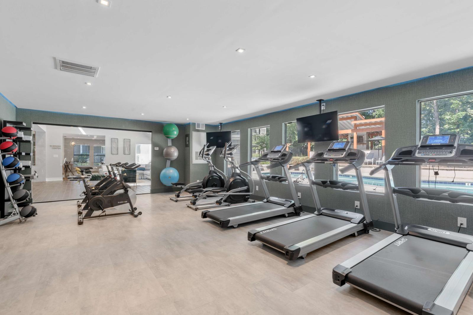 Modern fitness center with treadmills, ellipticals, stationary bikes, and free weights, featuring large windows overlooking the pool and outdoor lounge area.