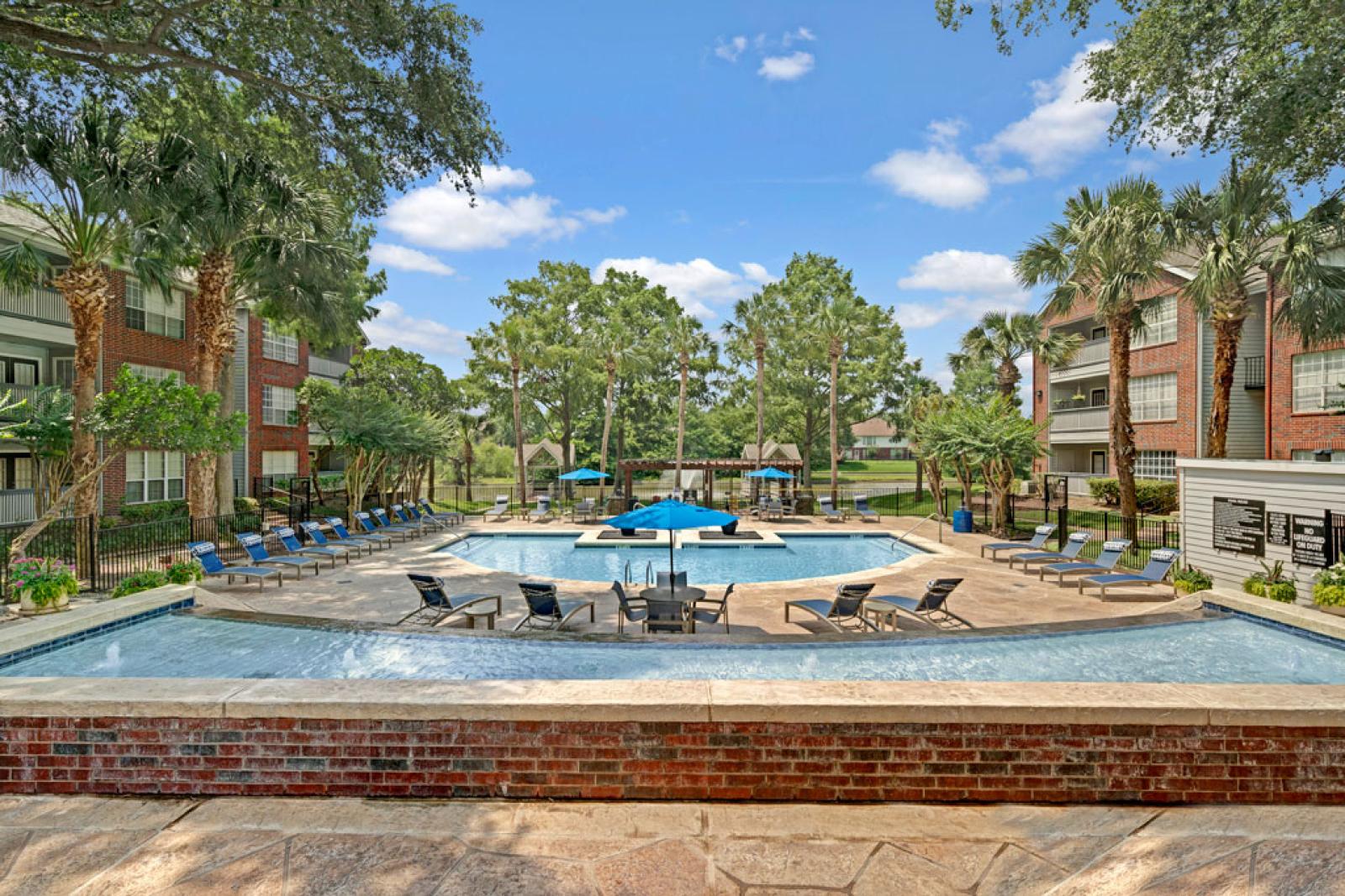 Resort-style swimming pool area surrounded by lounge chairs, palm trees, and red brick apartment buildings under a bright blue sky.