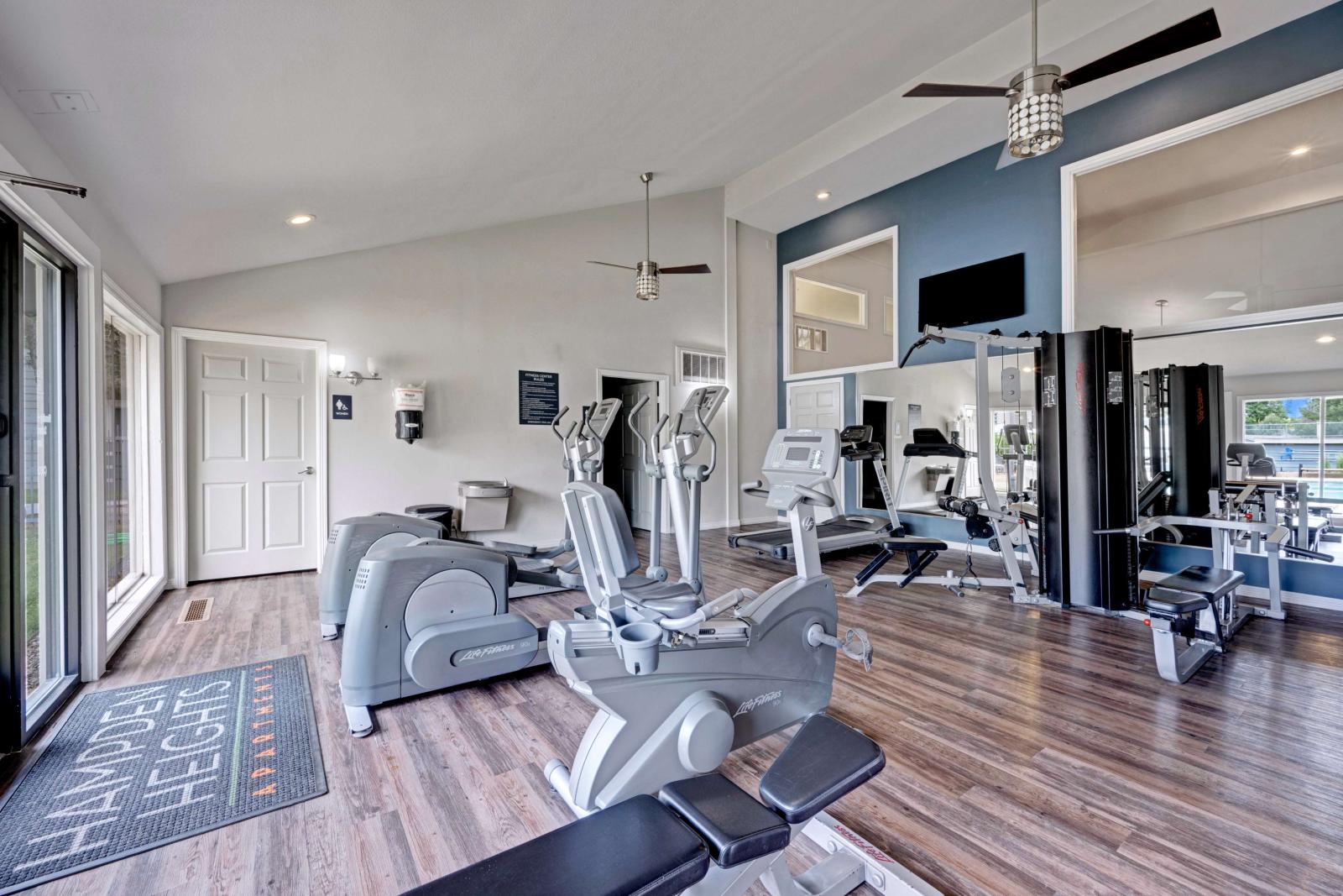 Modern fitness center with cardio and strength training equipment, large mirrors, wood-style flooring, and natural light from floor-to-ceiling windows.