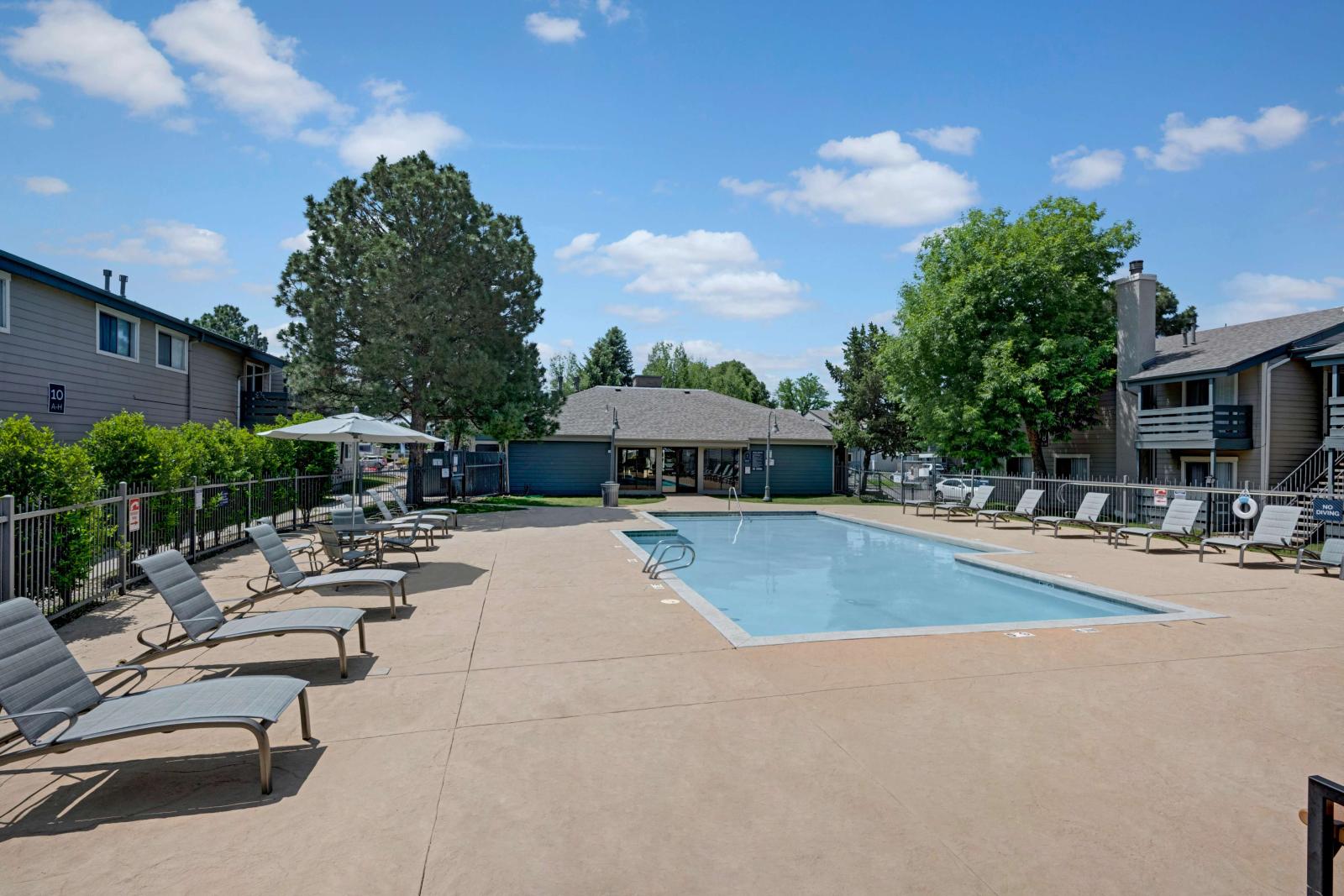 Outdoor swimming pool with surrounding lounge chairs, umbrellas, and fenced-in area, set in a residential apartment community with trees and blue skies.