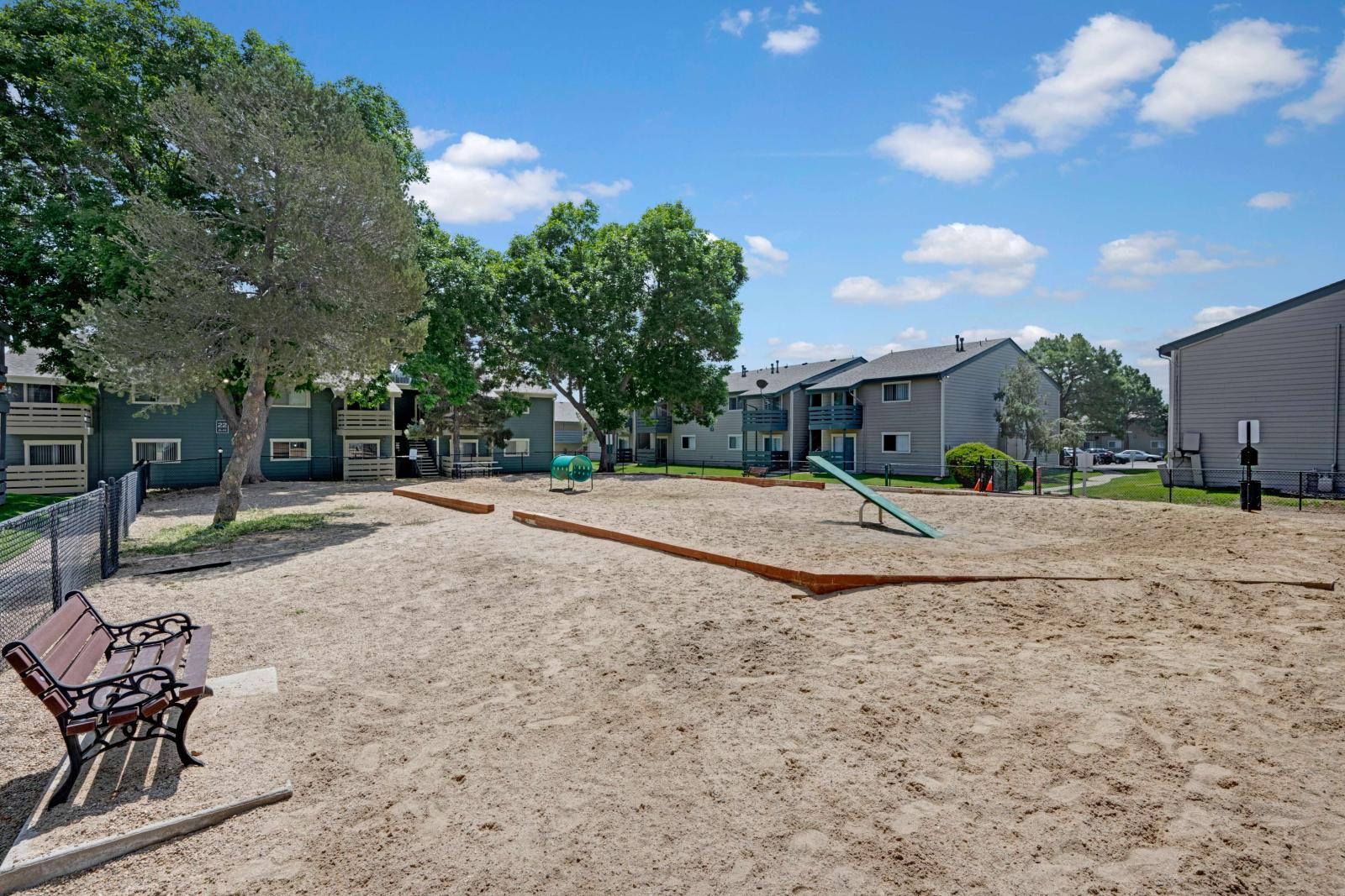 Outdoor dog park with sand surface, seesaw, and play tunnel, surrounded by mature trees and apartment buildings in the background.
