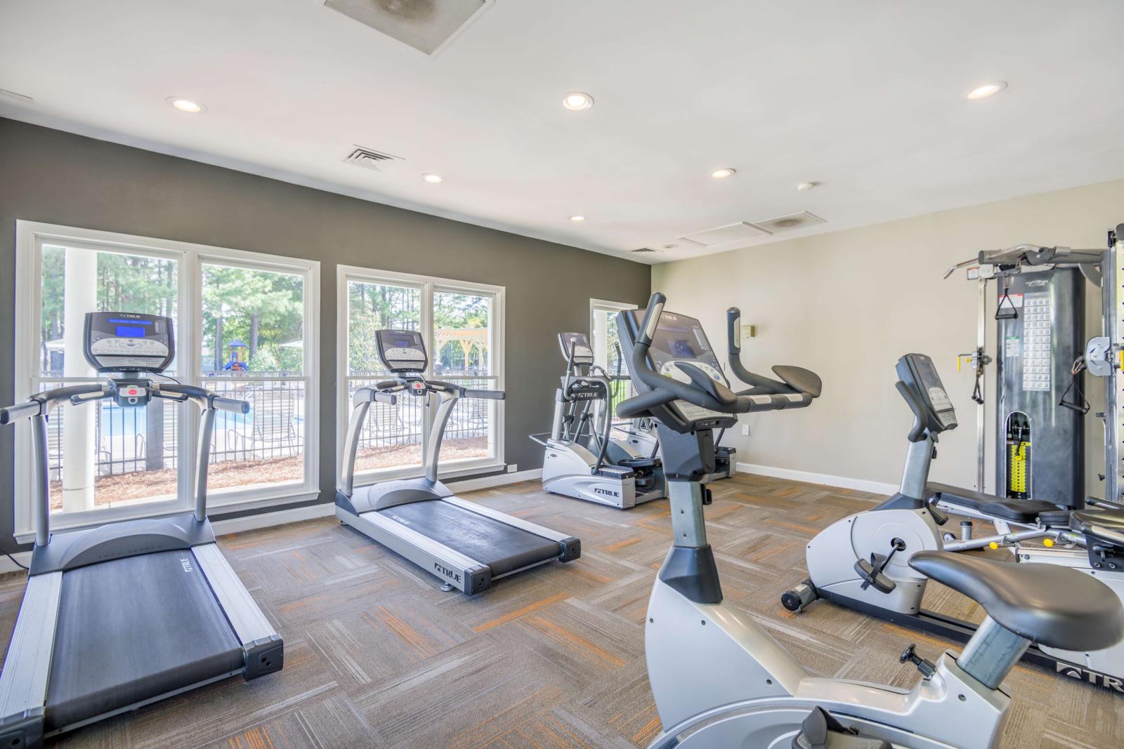  Bright fitness center with large windows, treadmills, ellipticals, stationary bikes, and strength training equipment on a patterned carpeted floor.