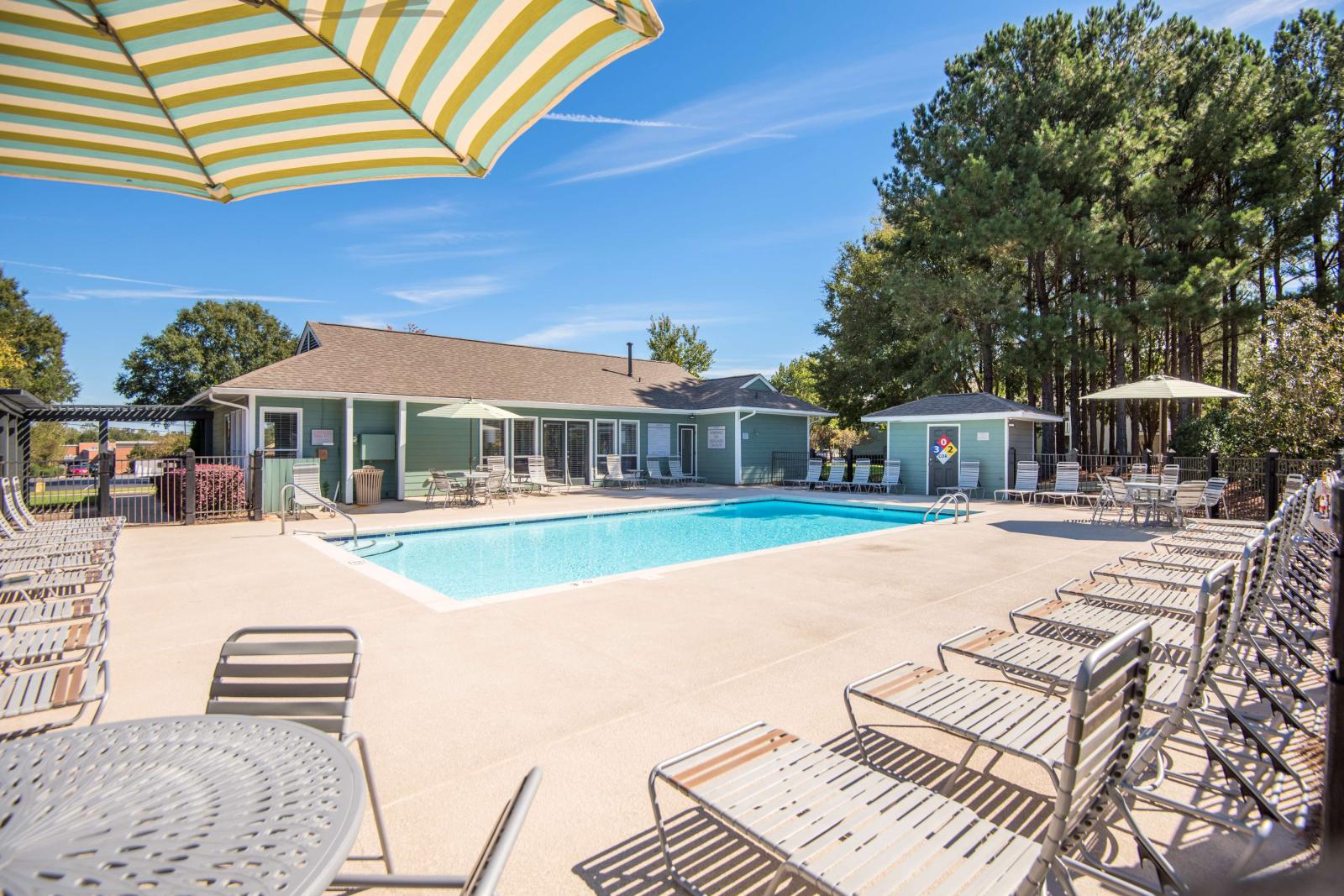 Outdoor swimming pool area with lounge chairs, patio tables, and umbrellas, surrounded by tall pine trees and a clubhouse building.