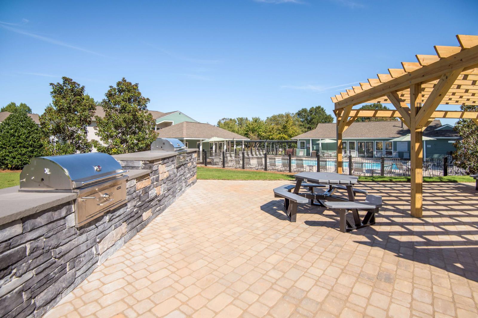 Outdoor grilling area with built-in stainless steel grills, stone countertops, picnic seating under a wooden pergola, and a view of the swimming pool and clubhouse in the background.