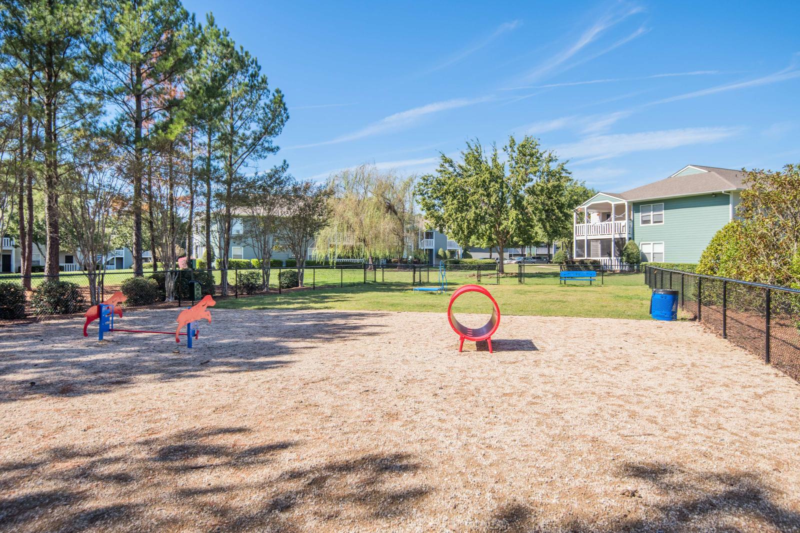 Fenced dog park with agility equipment including a red tunnel and spring-mounted dinosaur shapes, surrounded by trees, benches, and nearby apartment buildings.