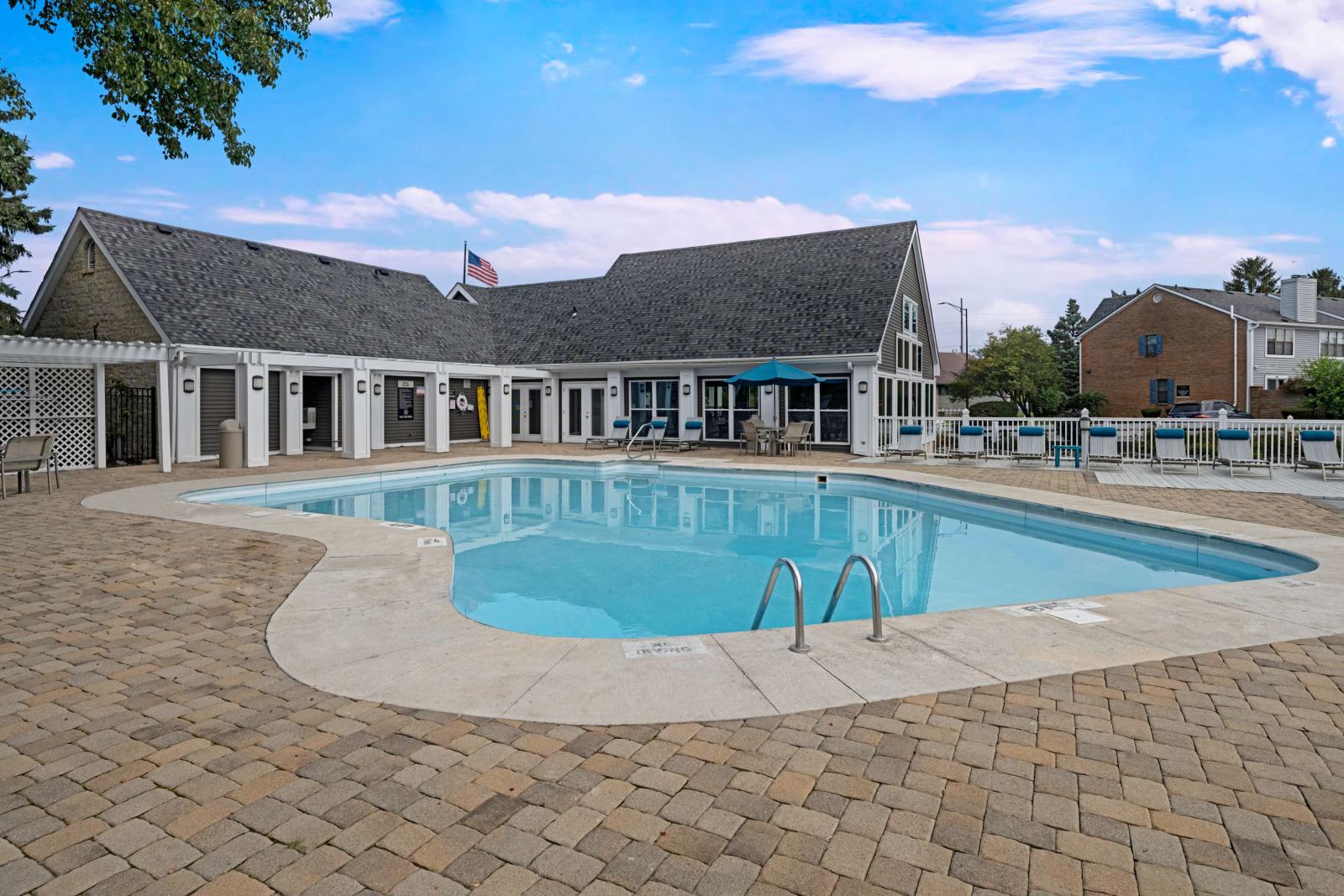 Inviting outdoor pool area with a curved swimming pool, cushioned lounge chairs, and a classic clubhouse building featuring white trim, gabled roofing, and a shaded patio with seating.