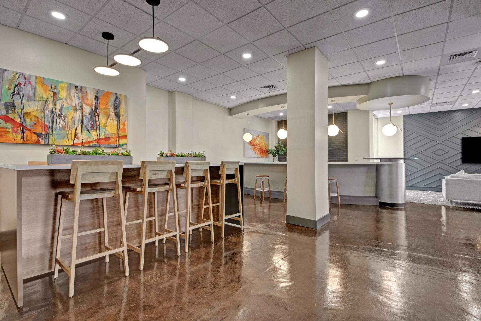 Modern resident lounge area featuring high-top seating with wooden barstools, pendant lighting, vibrant abstract artwork, and polished concrete floors.
