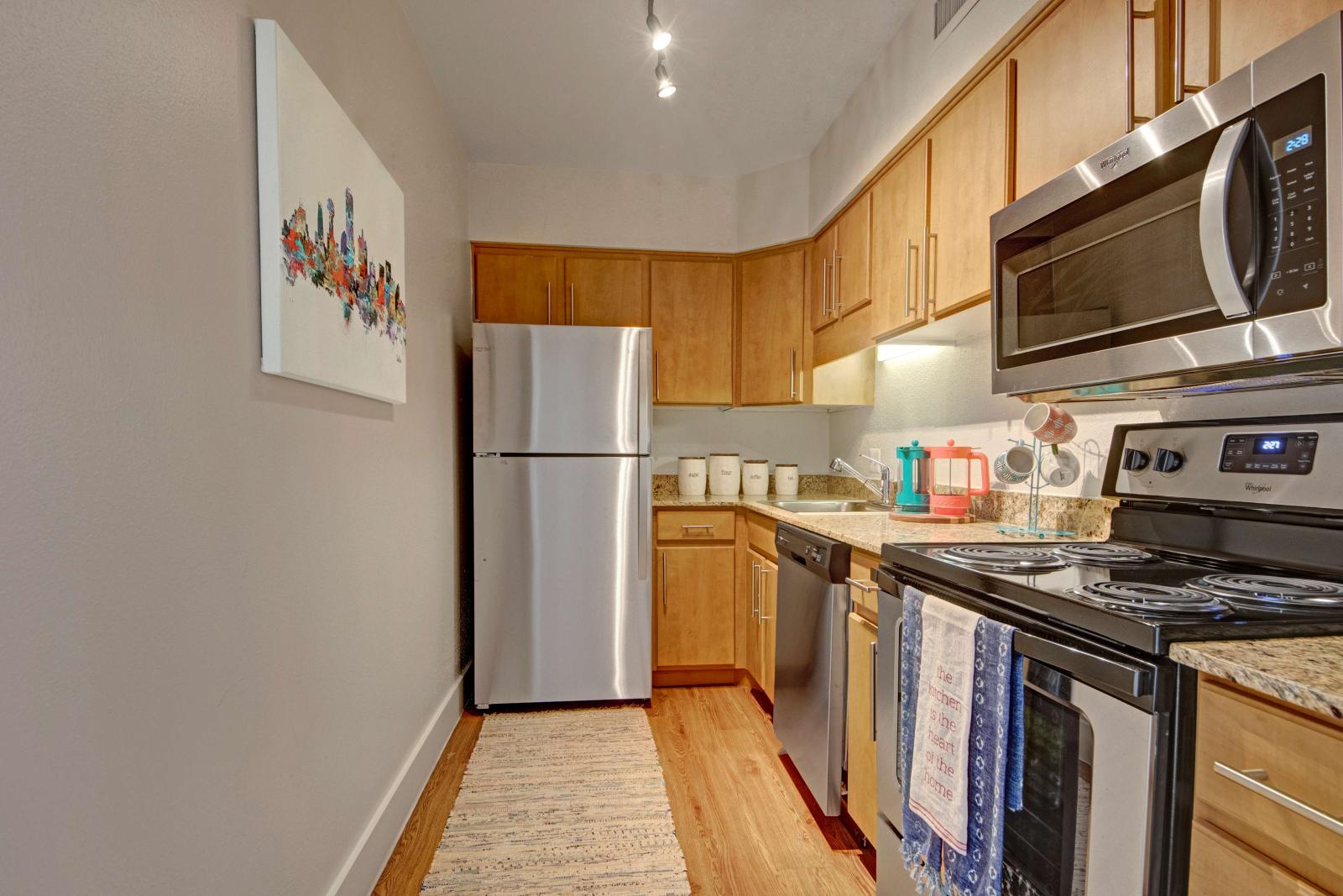 Contemporary galley kitchen with light wood cabinetry, stainless steel appliances, granite countertops, and a cozy decorative rug on wood-style flooring.