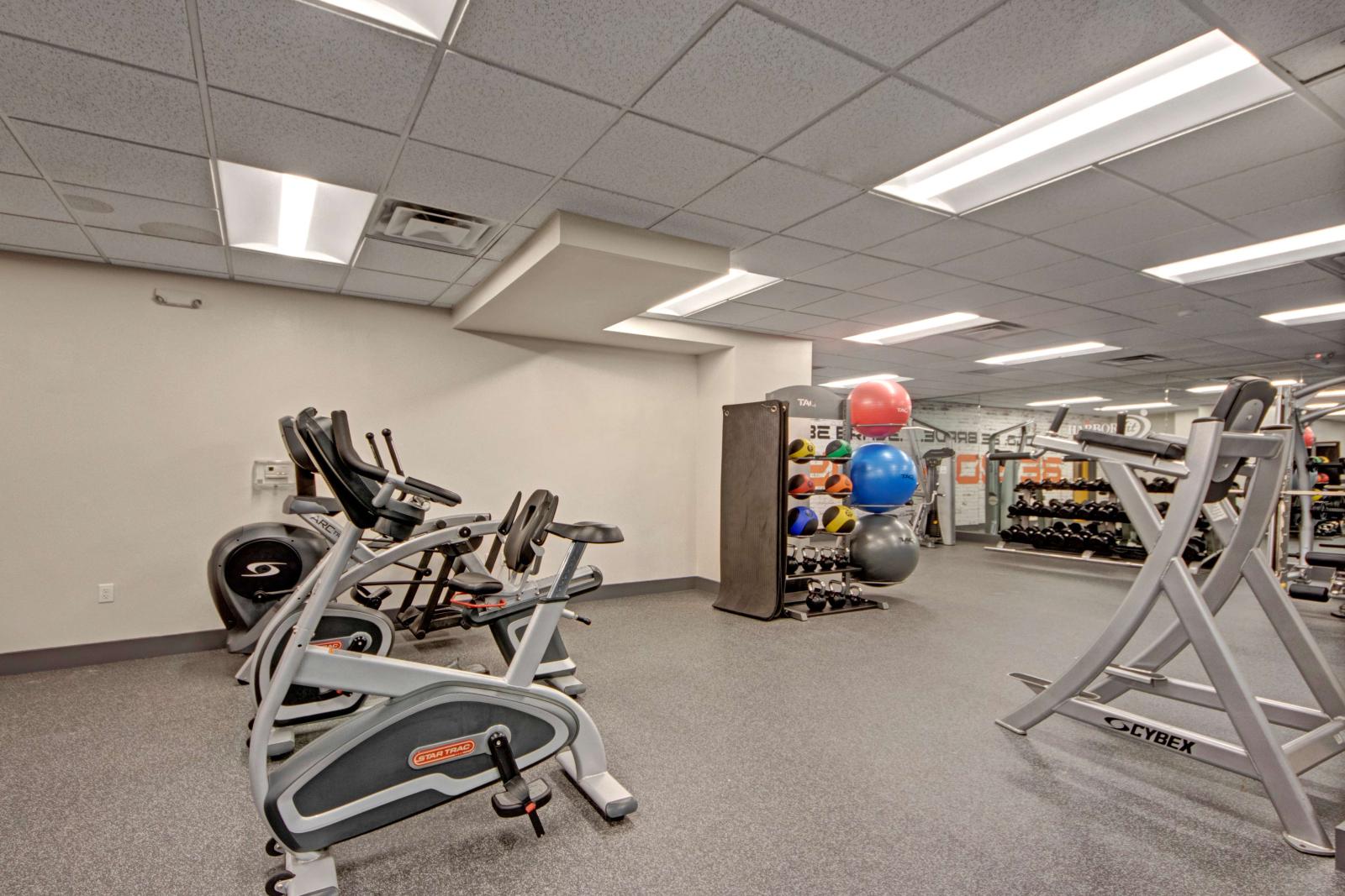 Spacious fitness center featuring cardio machines, free weights, resistance equipment, and colorful exercise balls in a brightly lit room with mirrored walls.